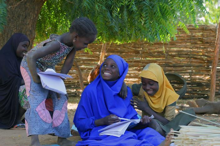 The daughters are smiling at each other while sitting outside in the shadow with their school books.