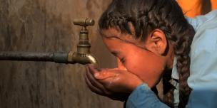 A girl in school dress and braids drinks water from a tap in the late afternoon sunlight