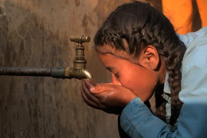 A girl in school dress and braids drinks water from a tap in the late afternoon sunlight