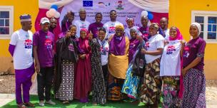 A diverse group of community members and organizers smiling together in front of an Oxfam International Women's Day 2026 banner in Kwaya Kusar, North-East Nigeria.