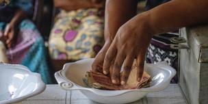 A close-up shot of a person's hands placing Nigerian currency notes into a white bowl on a table. This represents the weekly contributions of a VSLA group in Nigeria.