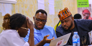 Three Oxfam colleagues lean in closely together to collaborate over a laptop screen during an intensive working session. Photo Credit: Maxwell Osarenkhoe/Oxfam