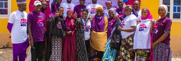 A diverse group of community members and organizers smiling together in front of an Oxfam International Women's Day 2026 banner in Kwaya Kusar, North-East Nigeria.