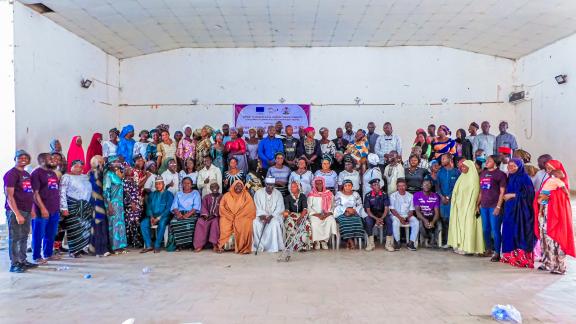 A large, diverse group of community members, traditional leaders, and Oxfam partners posing for a commemorative photo to celebrate International Women's Day 2026 under the Rights Justice Action theme inside a hall in Guyuk, North East Nigeria.