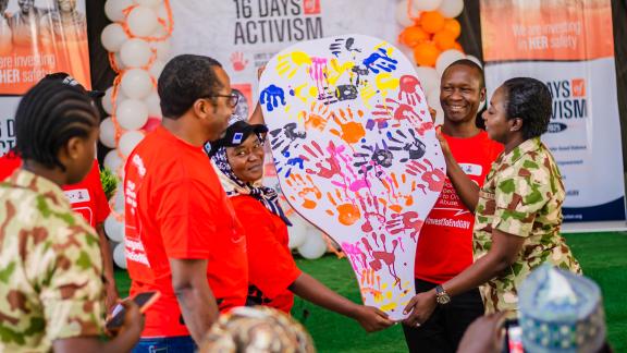 Oxfam team holding up a large, white, whistle-shaped board covered in diverse colored handprints, symbolizing a collective call to action against violence.