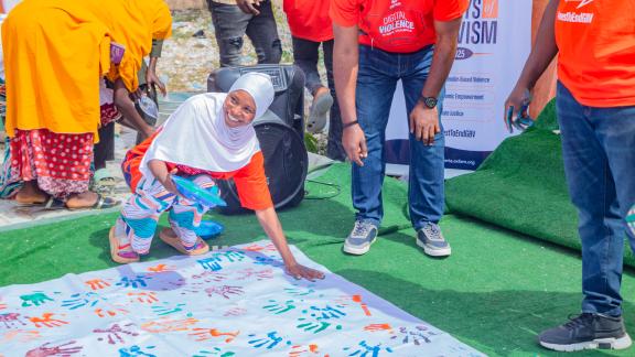 Student kneeling, placing a colorful handprint on a large white canvas on the ground as part of a collective commitment ceremony against GBV.