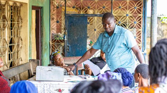 A male facilitator in a blue shirt speaks to a group of women during a VSLA meeting. An elderly woman is seated in the foreground.
