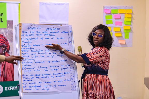 A female presenter holds a microphone and points to a flipchart titled 'FEMINIST APPROACH' to break down gender-transformative strategies. Photo Credit: Maxwell Osarenkhoe/Oxfam