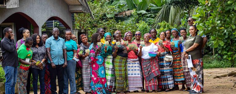 A large group of men and women, members of a Village Savings and Loan Association (VSLA) in Nigeria, stand together outside with Oxfam staff. The women are holding up their VSLA passbooks.