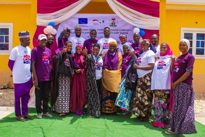 A diverse group of community members and organizers smiling together in front of an Oxfam International Women's Day 2026 banner in Kwaya Kusar, North-East Nigeria.