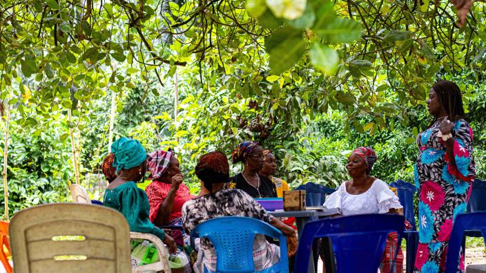 A group of women are gathered in a community under a canopy of trees, sitting around a table for a VSLA meeting.