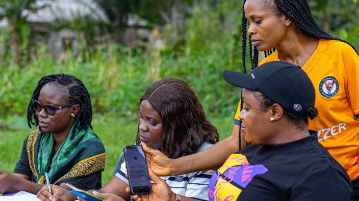 A group of women gathered outside, with one woman holding up a smartphone showing a mobile application to another. This illustrates the use of digital tools for VSLA record-keeping.