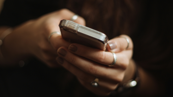 A close-up shot shows a person's hands holding and using a smartphone, with several rings on their fingers.