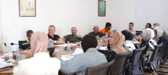 A group of people are gathered around a table, smiling and talking, during a digital rights training held by MIFTAH in Palestine.