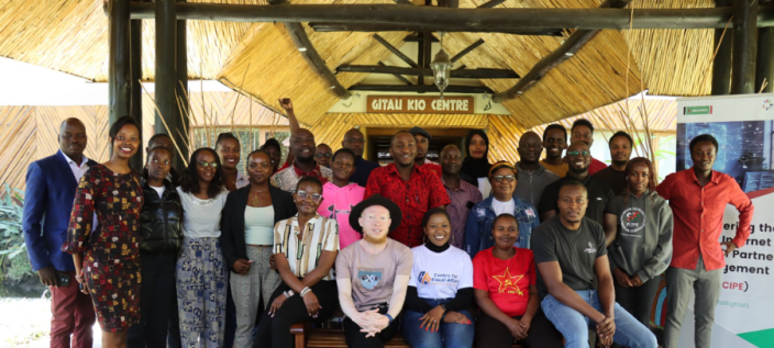 A group of people posing for a photo in front of a building with a thatched roof. The sign above them reads "GITAU KIO CENTRE".